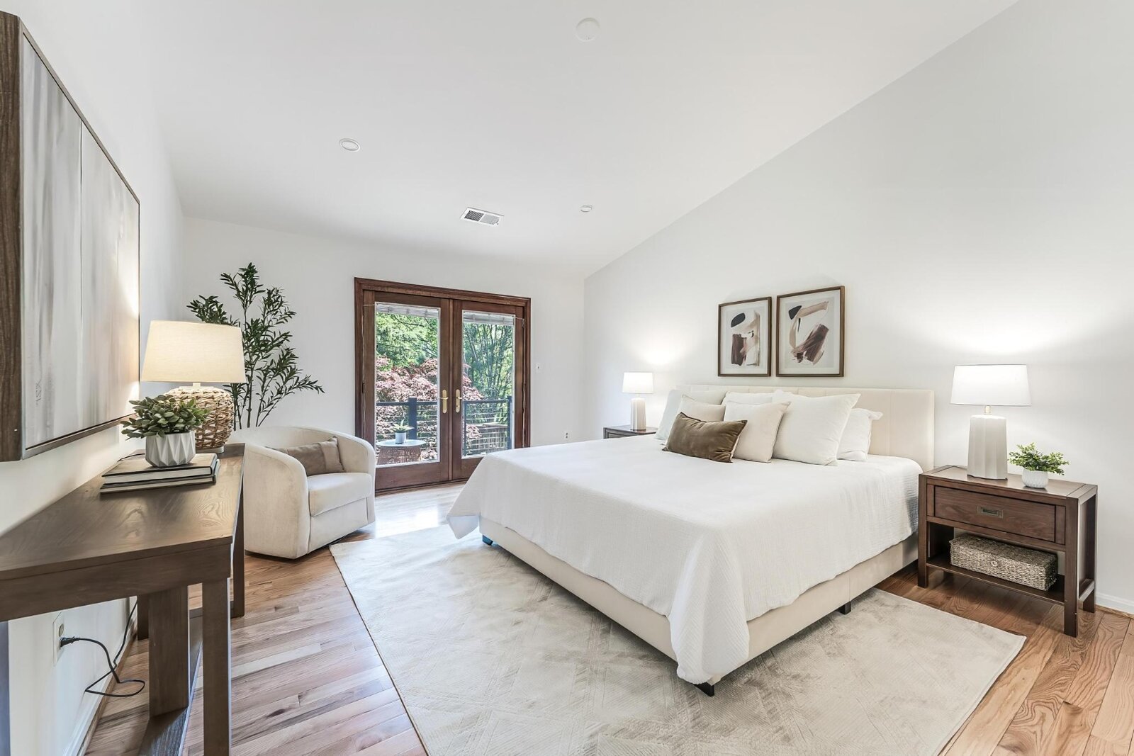 An elegantly staged primary bedroom with a white upholstered bed, warm wood nightstands, framed abstract art, and French doors opening to a balcony